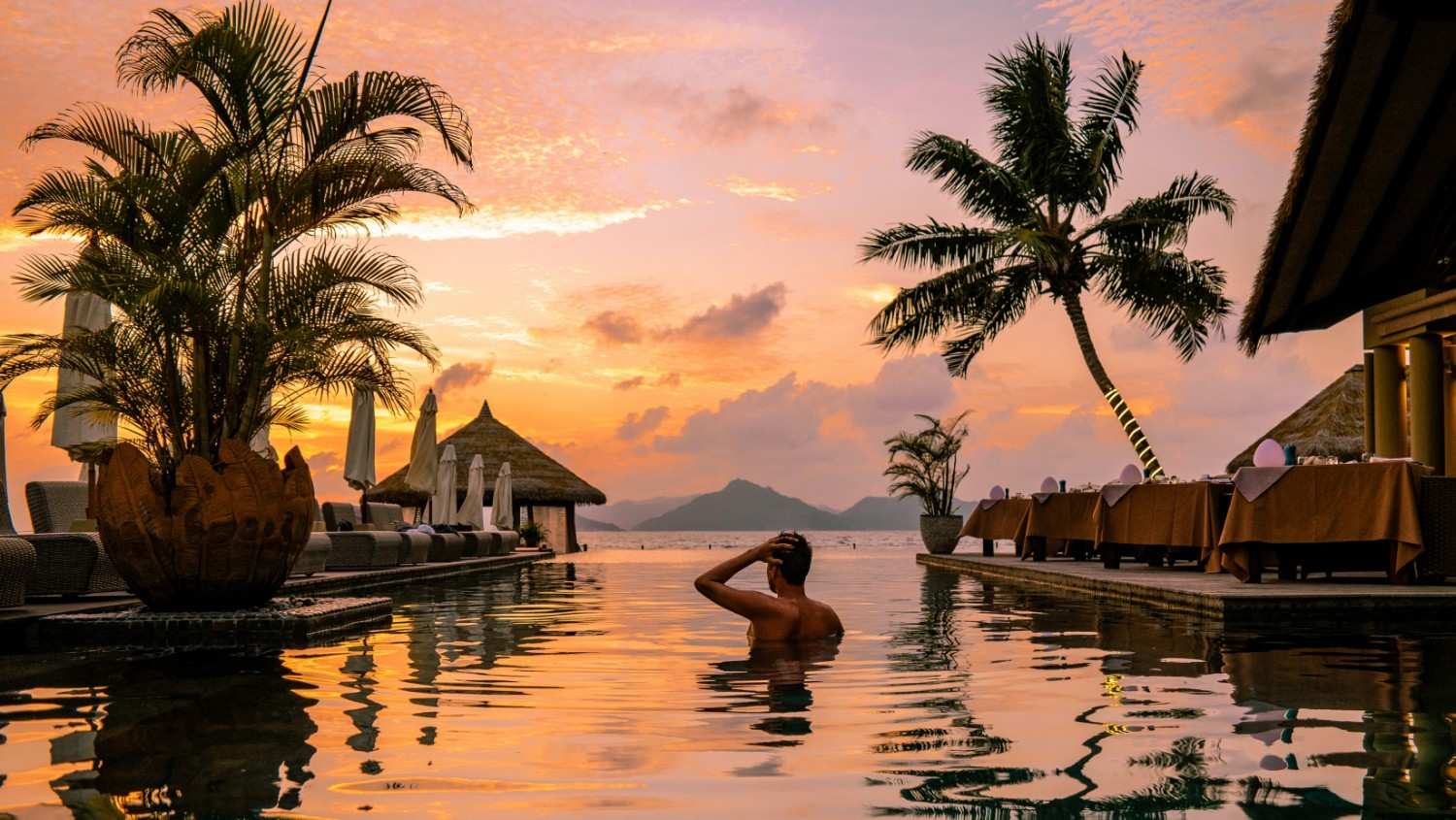 A woman stands in an infinity pool at sunset, looking out over a calm ocean with distant islands. Palm trees silhouette against the vibrant orange and pink sky, and tables are set for dining on a patio next to the pool.