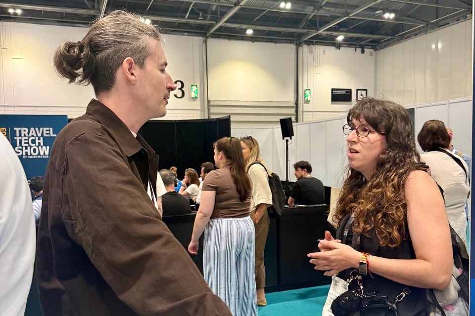 A man speaks to a woman wearing a lanyard at an exhibition booth with a coffee cup nearby.