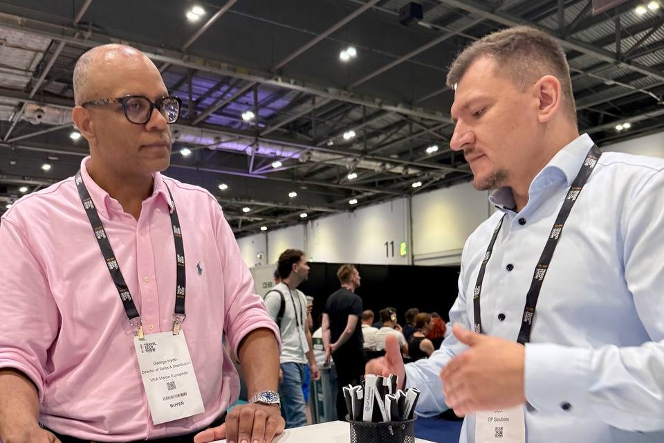 Two men having a conversation at the GP Solutions exhibition booth, standing across a small white table.
