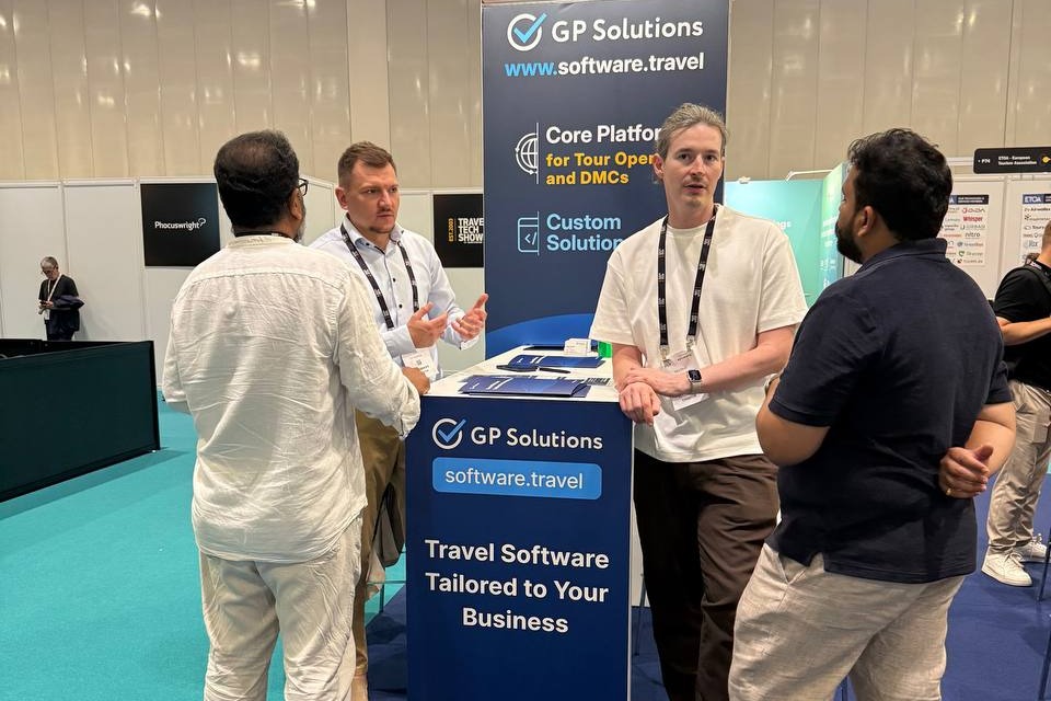 Three men talk at a GP Solutions trade‑show booth with a blue counter describing travel software tailored to your business behind them.