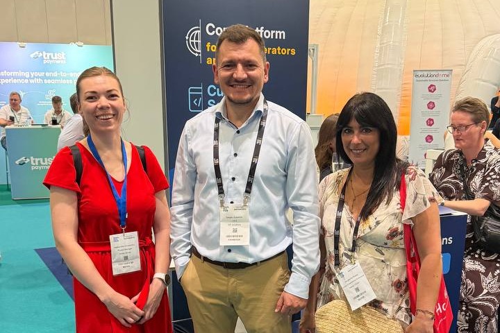 Three conference attendees posing for a photo at a trade show booth, smiling and wearing name badges.