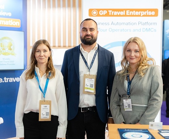 Three conference attendees stand at a GP Travel Enterprise booth, wearing name badges and smiling at the camera.